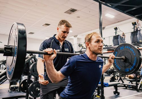 man in gym with gym instructor performing a barbell squat